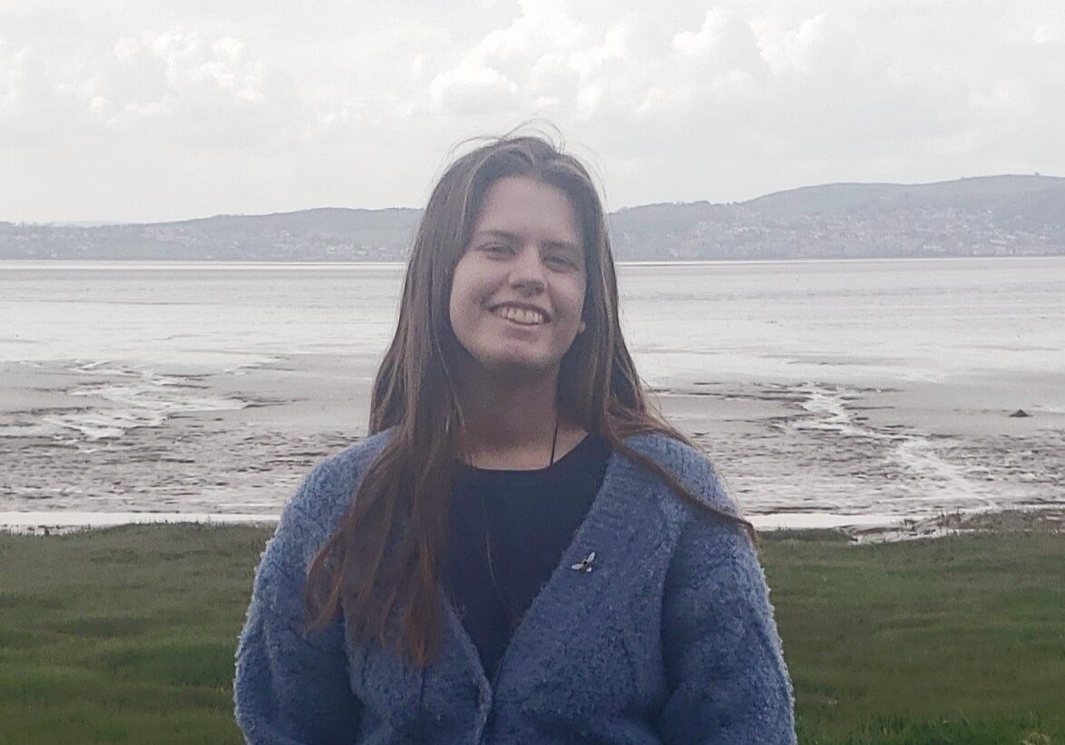 eleanor_23 Eleanor smiling broadly to camera with the beach and a sea behind her